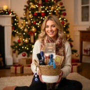 Smiling blonde woman holding a luxury Greek gift basket in a cozy, festive Christmas setting with lights and wrapped presents.